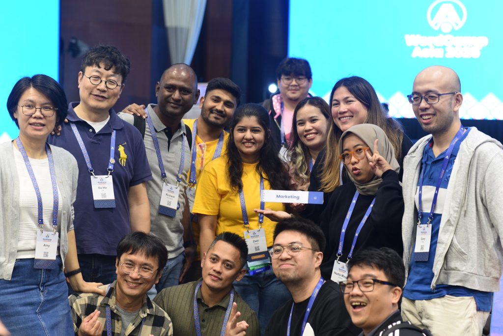 A diverse group of WordCamp Asia attendees posing together and smiling at the Marketing table during Contributor Day.