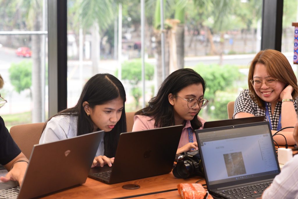 Three women focused on their laptops during a Contributor Day session, collaborating near large windows with natural light.