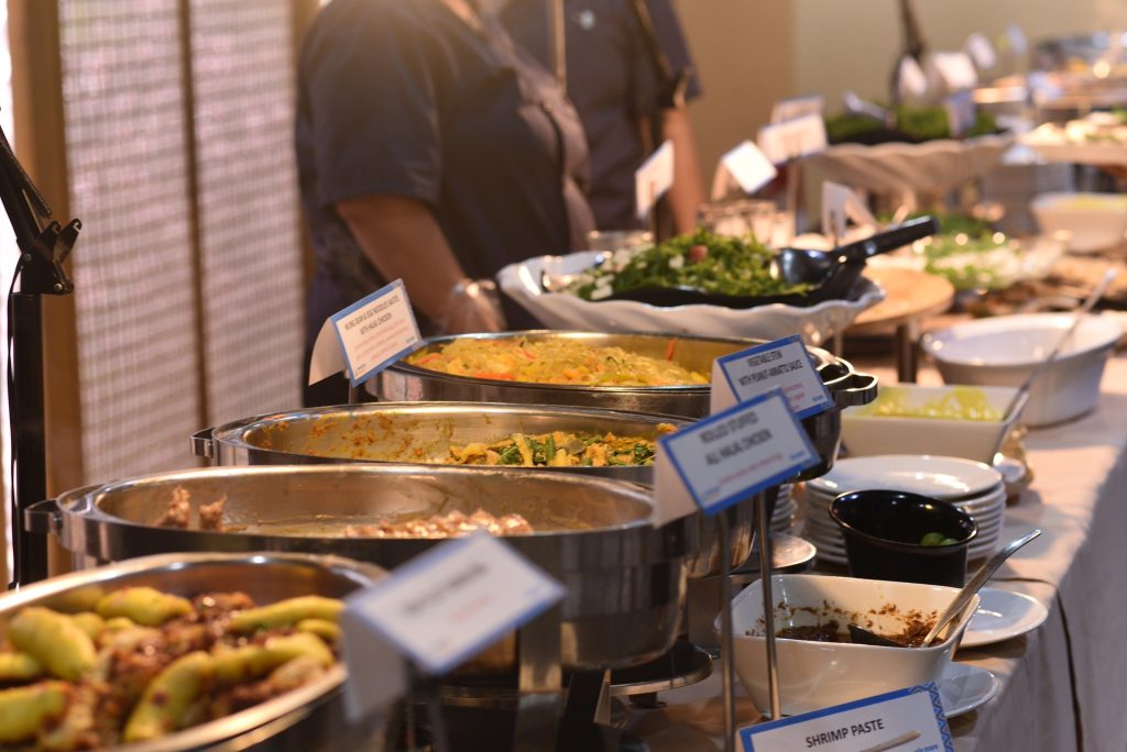 A buffet table at WordCamp Asia featuring a variety of dishes in stainless steel chafing trays, salads, condiments, and labeled food options prepared for attendees.